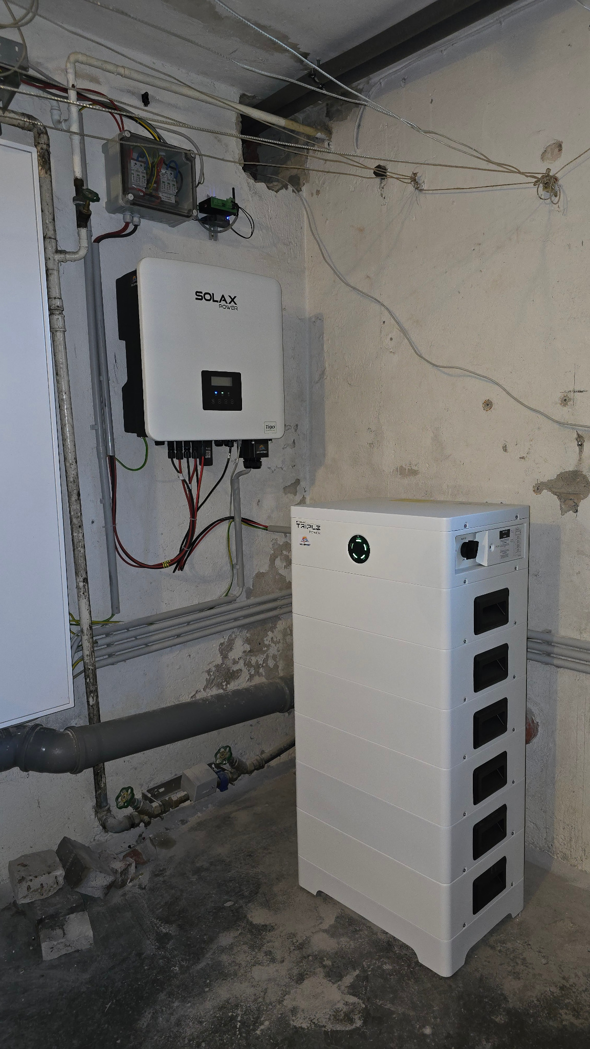 Biały inwerter Solax Power i akumulator energii na tle surowej ściany w piwnicy. Widoczne przewody i instalacje. Realizacja instalacji fotowoltaicznej.