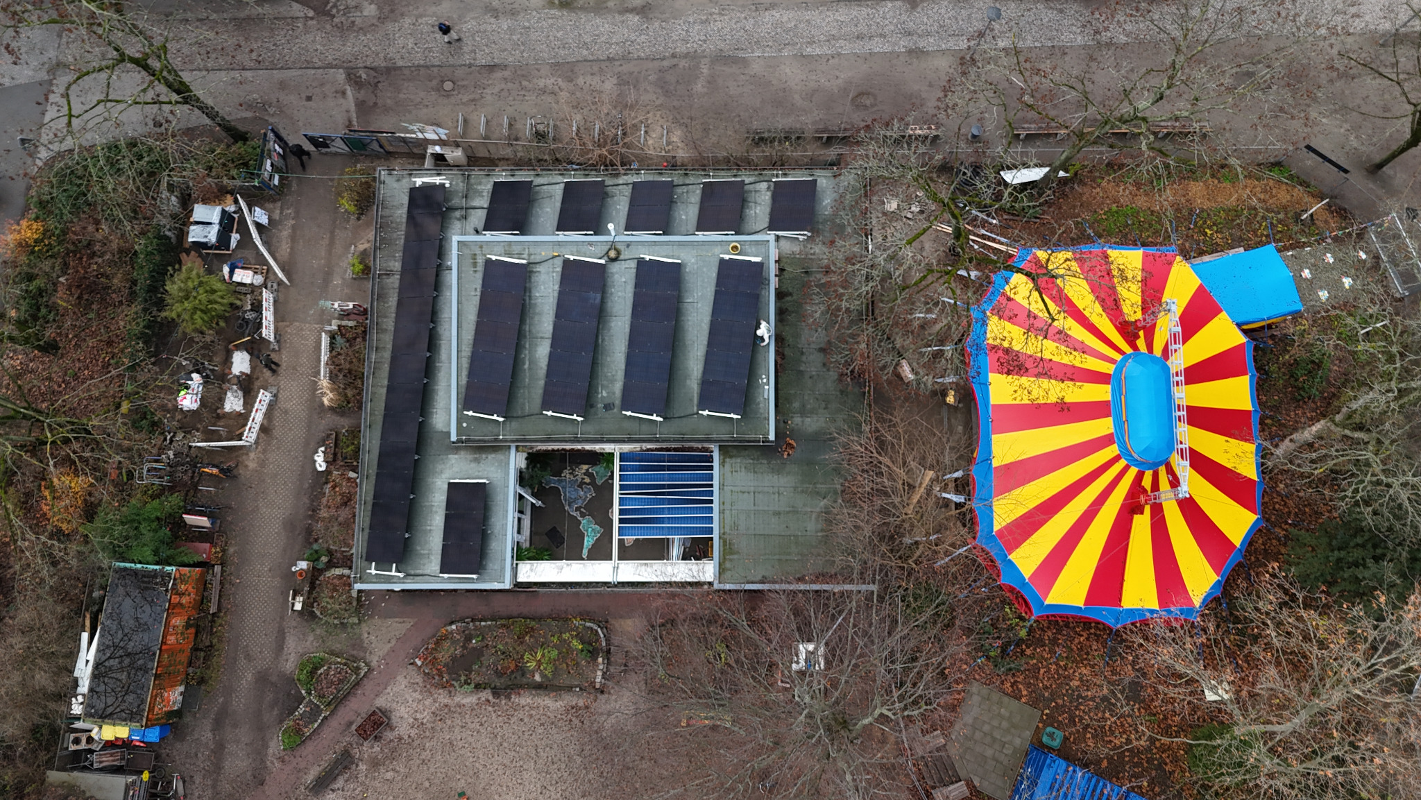 Widok z góry na budynek z panelami słonecznymi i kolorowy namiot cyrkowy. Nietypowe sąsiedztwo instalacji fotowoltaicznej i atrakcji rozrywkowej.