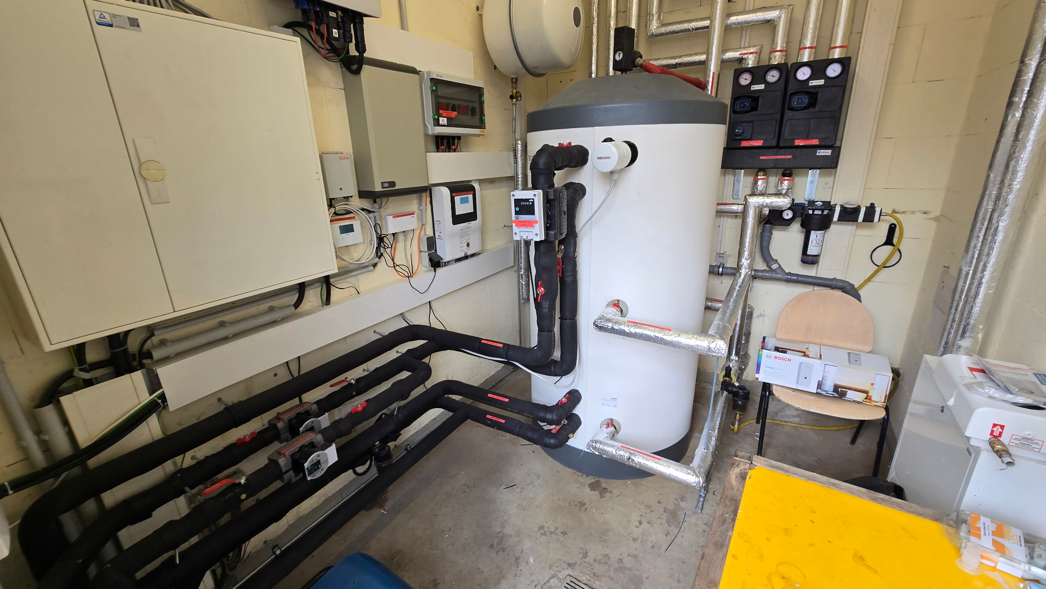 Węzeł cieplny z zbiornikiem akumulacyjnym, rurami w izolacji i osprzętem. Widoczne skrzynki elektryczne i sterowniki. Elementy instalacji grzewczej w kotłowni.