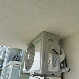 KlimatFix - Zamontowana jednostka zewnętrzna klimatyzacji Aerostar na balkonie budynku mieszkalnego. Widoczne rury i przewody. Ujęcie z dołu, z widokiem na sufit balkonu.