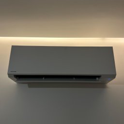 KlimatFix - Szary klimatyzator Toshiba na ścianie, podświetlony od góry listwą LED. Minimalistyczny design, widoczne logo i niebieska dioda. Ujęcie centralne, równomierne oświetlenie.