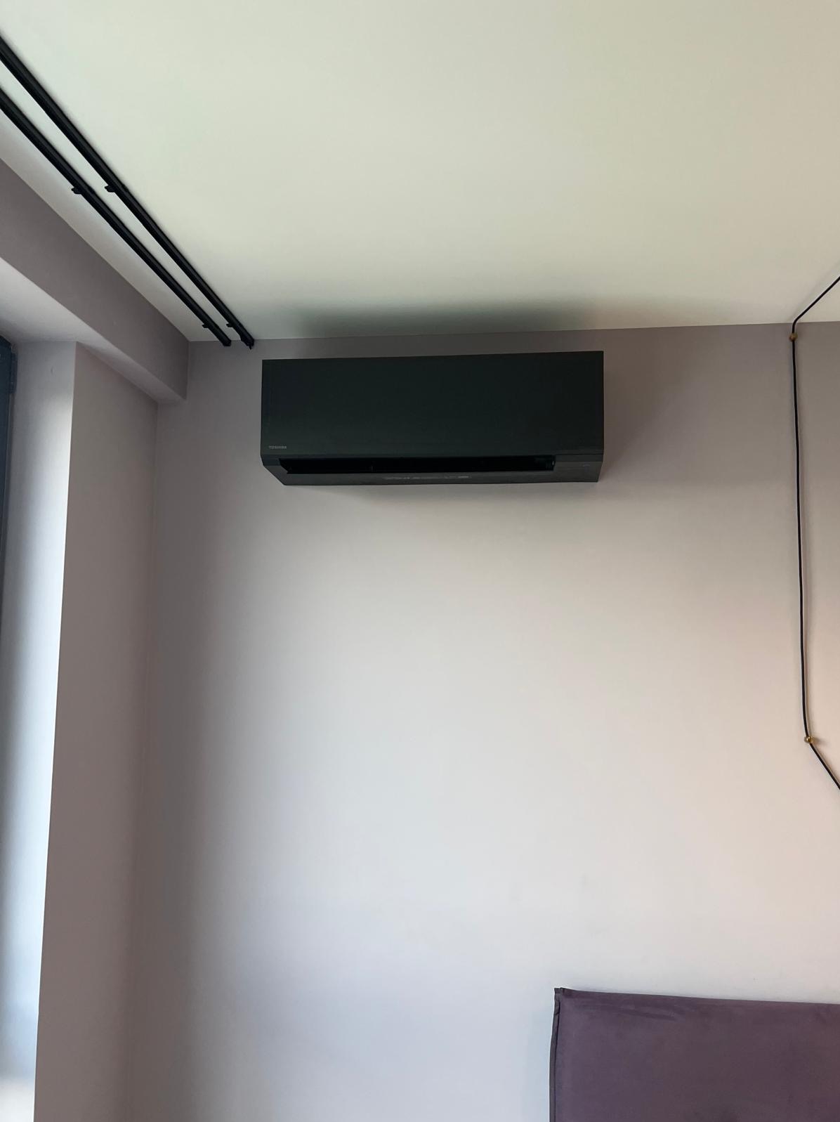 Czarny klimatyzator Toshiba zamontowany na ścianie w pokoju w Warszawie. Widoczne czarne szyny sufitowe i fragment wezgłowia łóżka. Ściana w odcieniach fioletu.