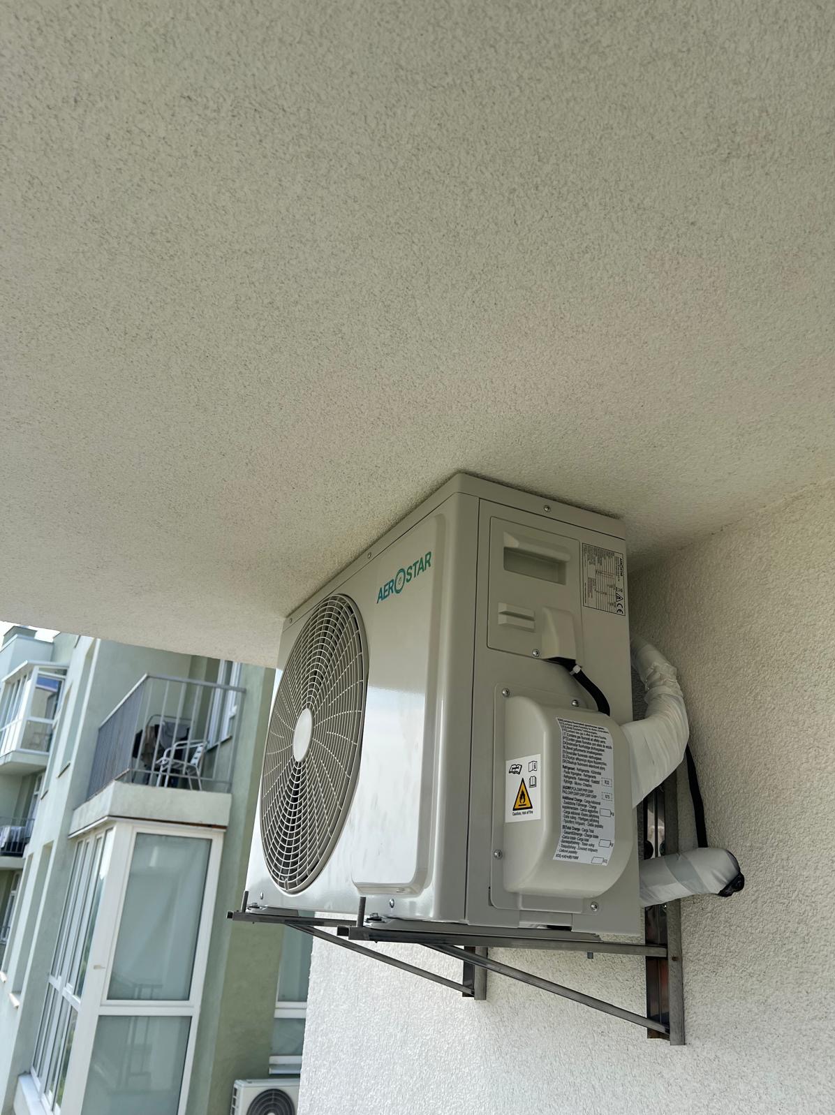 Zamontowana jednostka zewnętrzna klimatyzacji Aerostar na balkonie budynku mieszkalnego. Widoczne rury i przewody. Ujęcie z dołu, z widokiem na sufit balkonu.