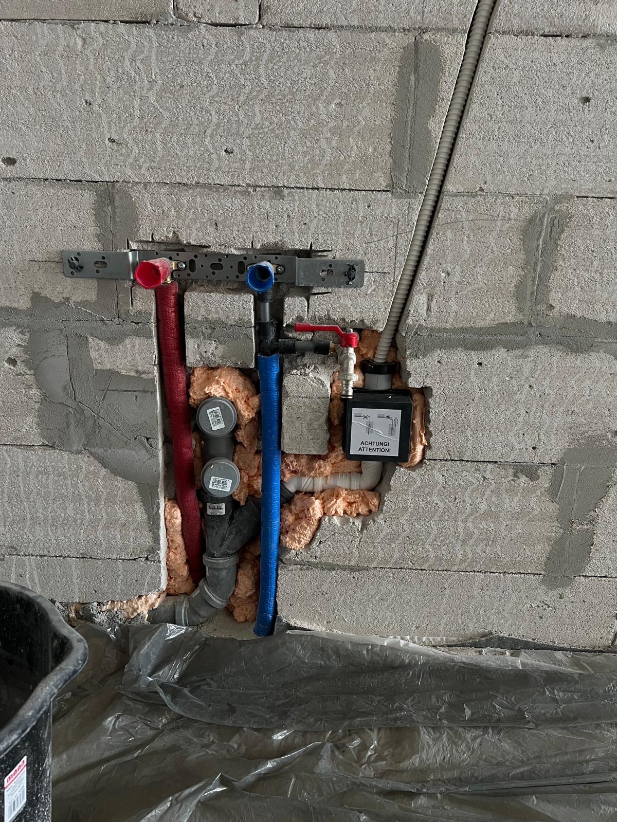Instalacja wodno-kanalizacyjna w surowym murze z widocznymi rurami (czerwona, niebieska, szara), pianką montażową i elementami mocującymi. Widoczna etykieta ostrzegawcza.
