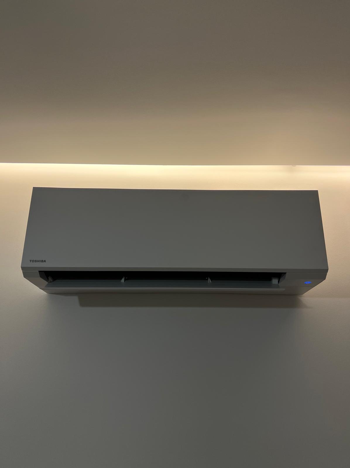 Szary klimatyzator Toshiba na ścianie, podświetlony od góry listwą LED. Minimalistyczny design, widoczne logo i niebieska dioda. Ujęcie centralne, równomierne oświetlenie.