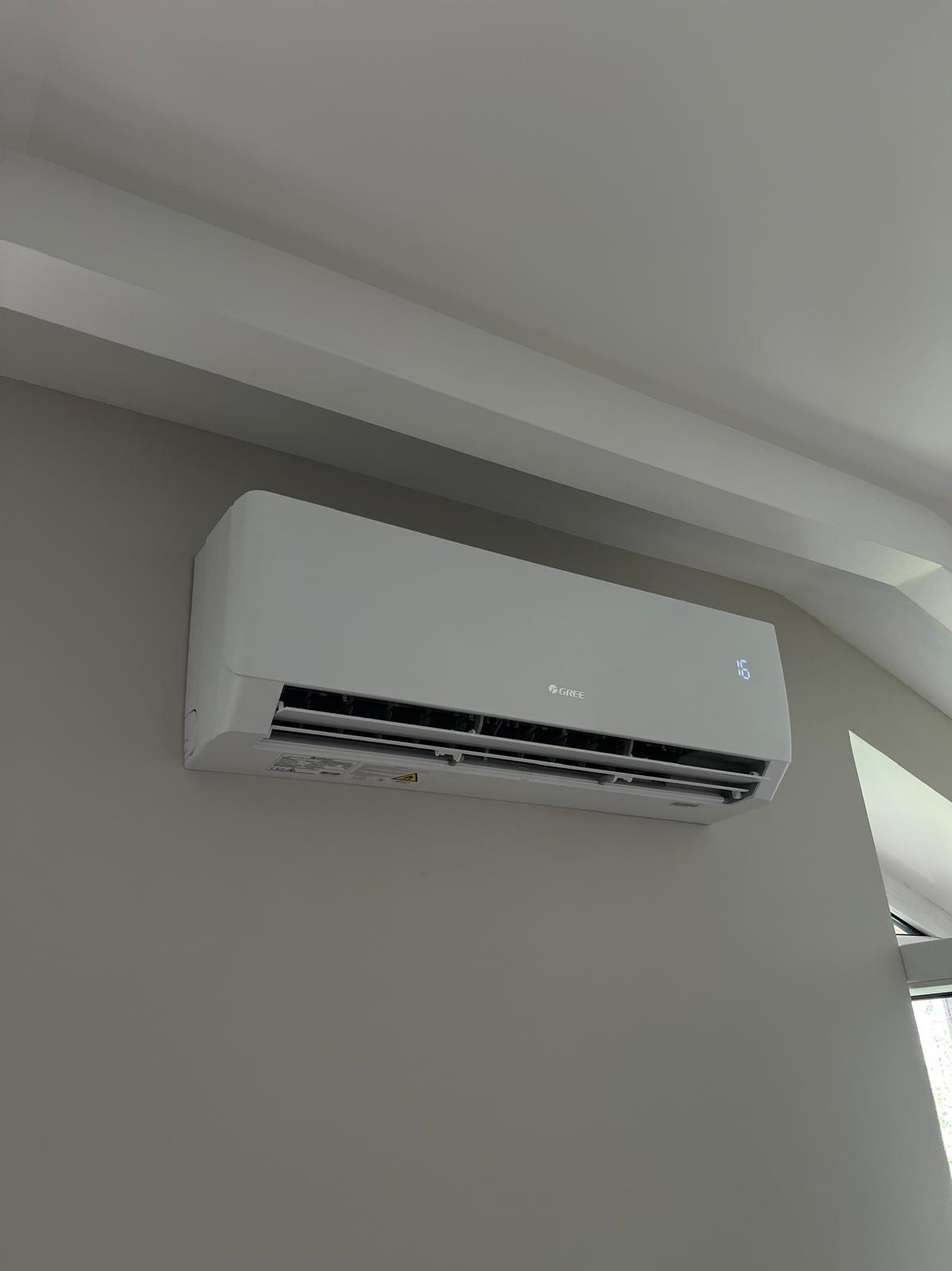 Biała klimatyzacja Gree zamontowana na szarej ścianie w pomieszczeniu z białym sufitem i listwami. Widoczny wyświetlacz temperatury.