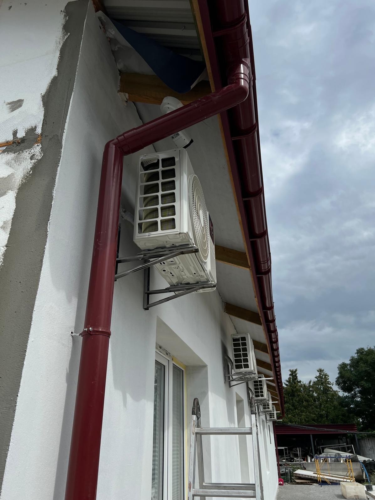 Biała elewacja budynku z zamontowanymi klimatyzatorami zewnętrznymi, widoczne rynny w kolorze bordo i drabina oparta o ścianę. Montaż klimatyzacji.