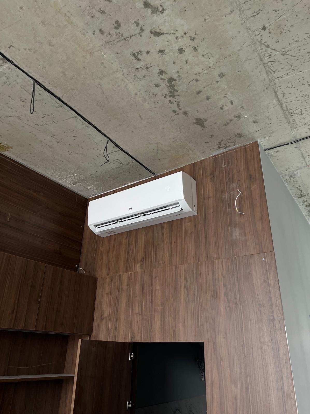 Biały klimatyzator ścienny zamontowany na brązowej, drewnianej ścianie w pomieszczeniu z surowym, betonowym sufitem i widocznymi kablami.