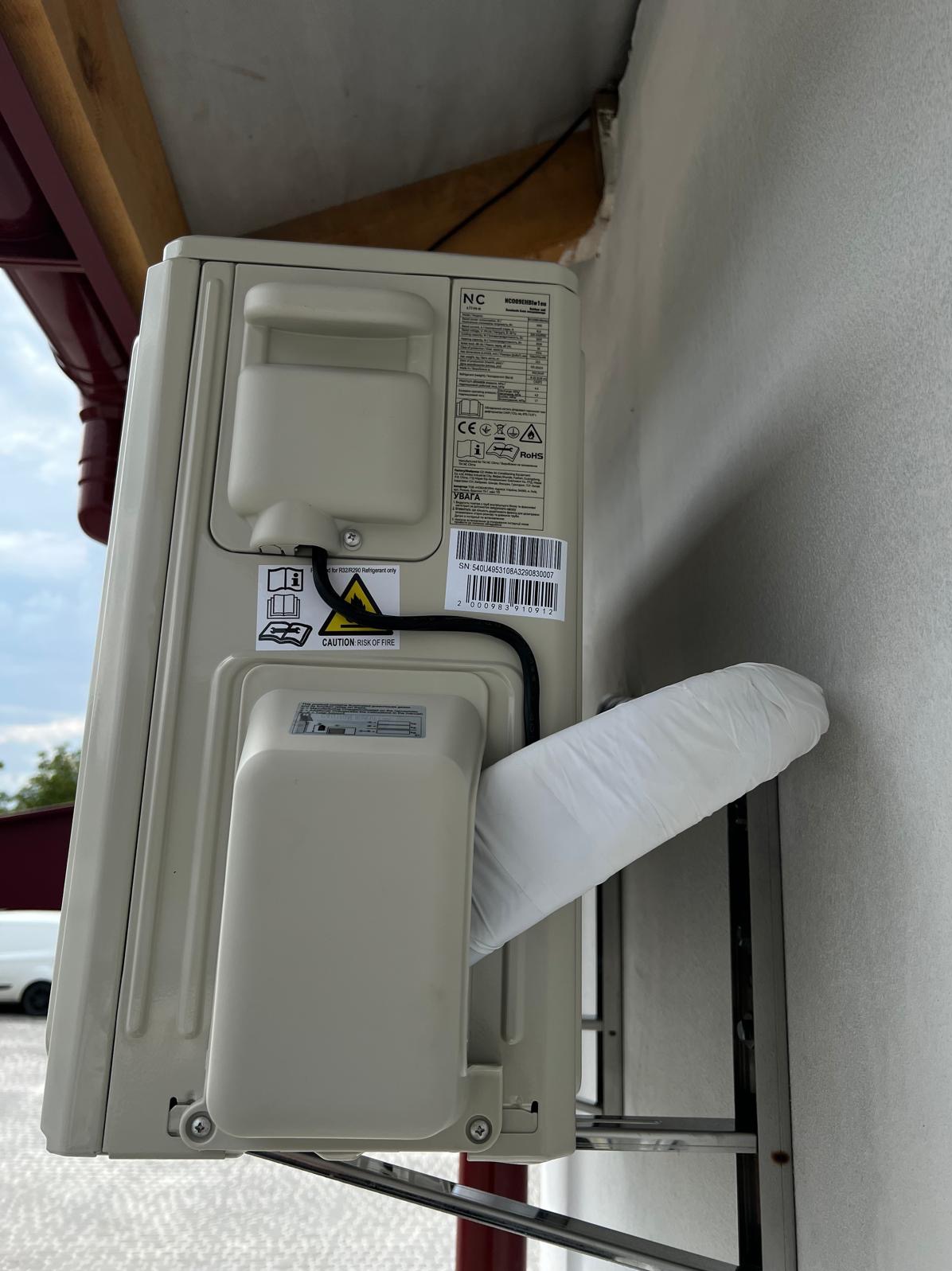 Zamontowana jednostka zewnętrzna klimatyzacji na metalowym stelażu, z widoczną etykietą informacyjną i rurą odprowadzającą skropliny owiniętą białą folią.