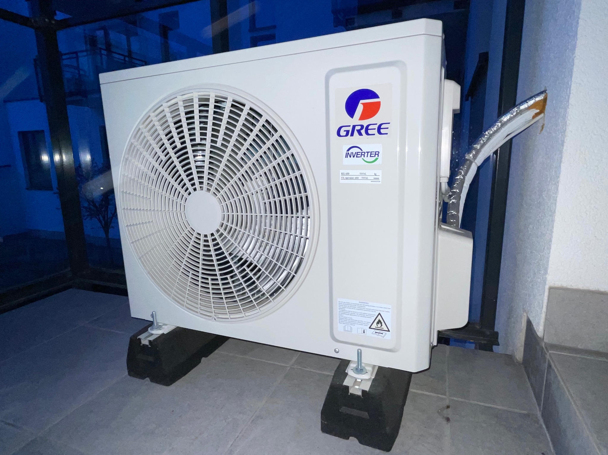 Biała jednostka zewnętrzna klimatyzacji GREE Inverter, zamontowana na czarnych podkładkach, z widocznymi przewodami instalacyjnymi na tle balkonu.