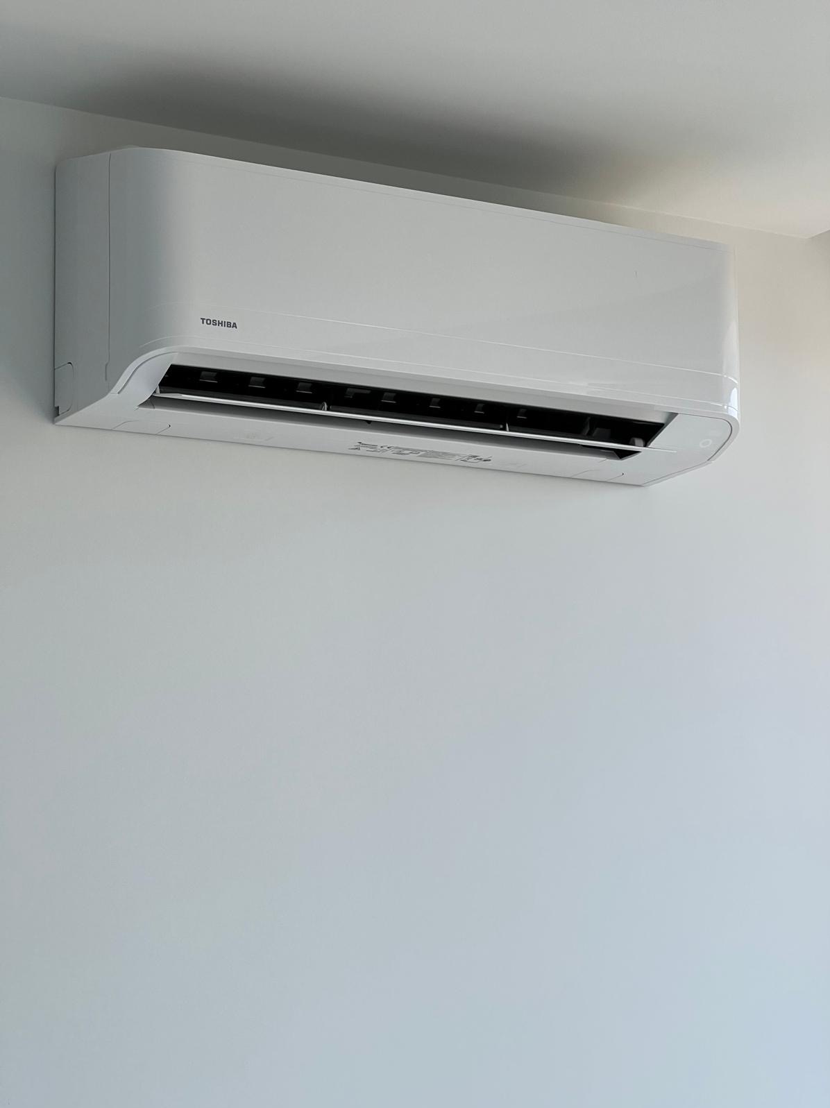 Biała klimatyzacja Toshiba zamontowana na jasnej ścianie. Minimalistyczny design, widoczne logo producenta. Ujęcie z dołu, podkreślające nowoczesny wygląd urządzenia.