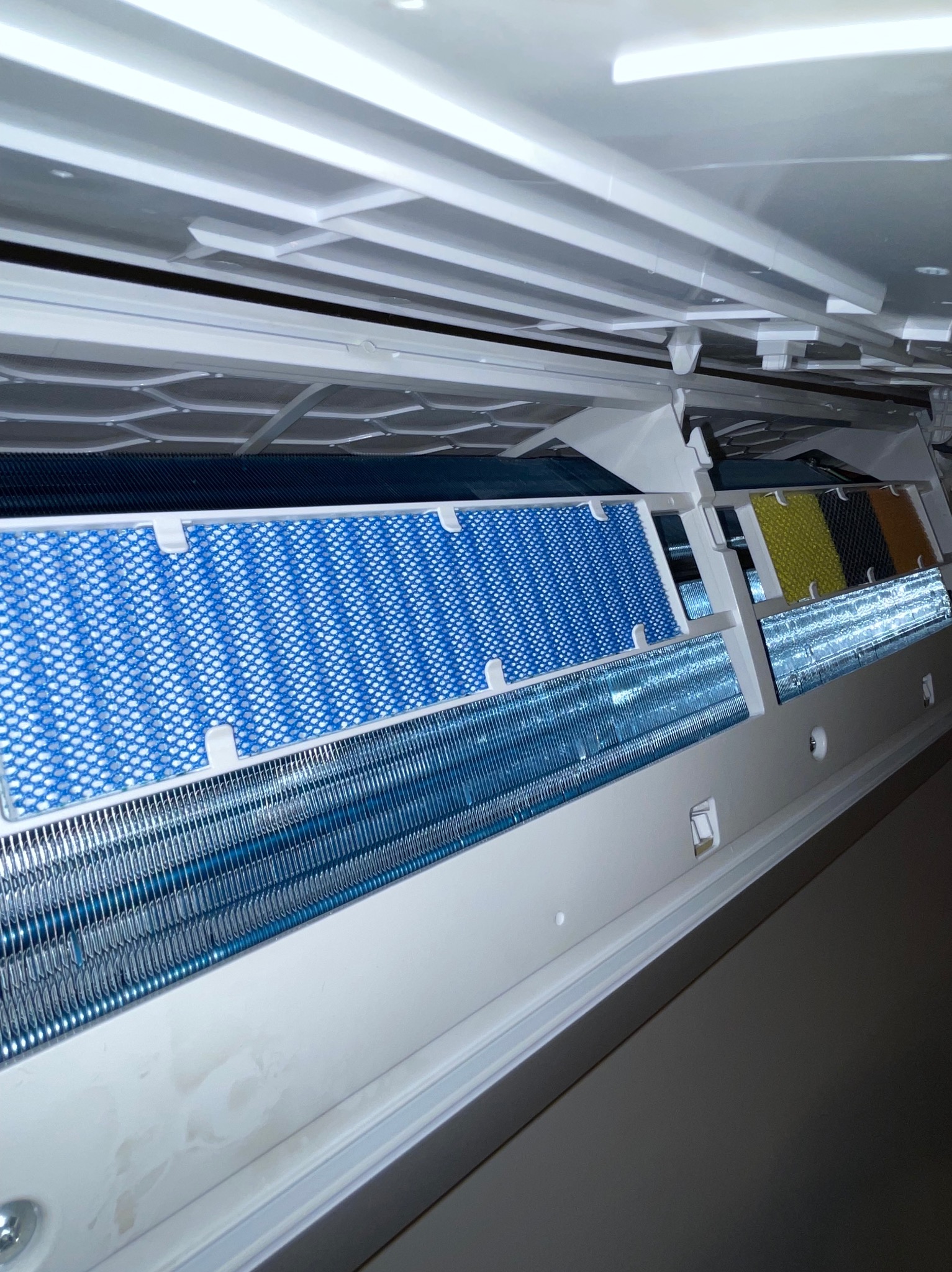 Wnętrze klimatyzatora z widocznymi filtrami (niebieski, żółty, czarny) i metalowymi lamelami. Widok od dołu, oświetlenie górne.