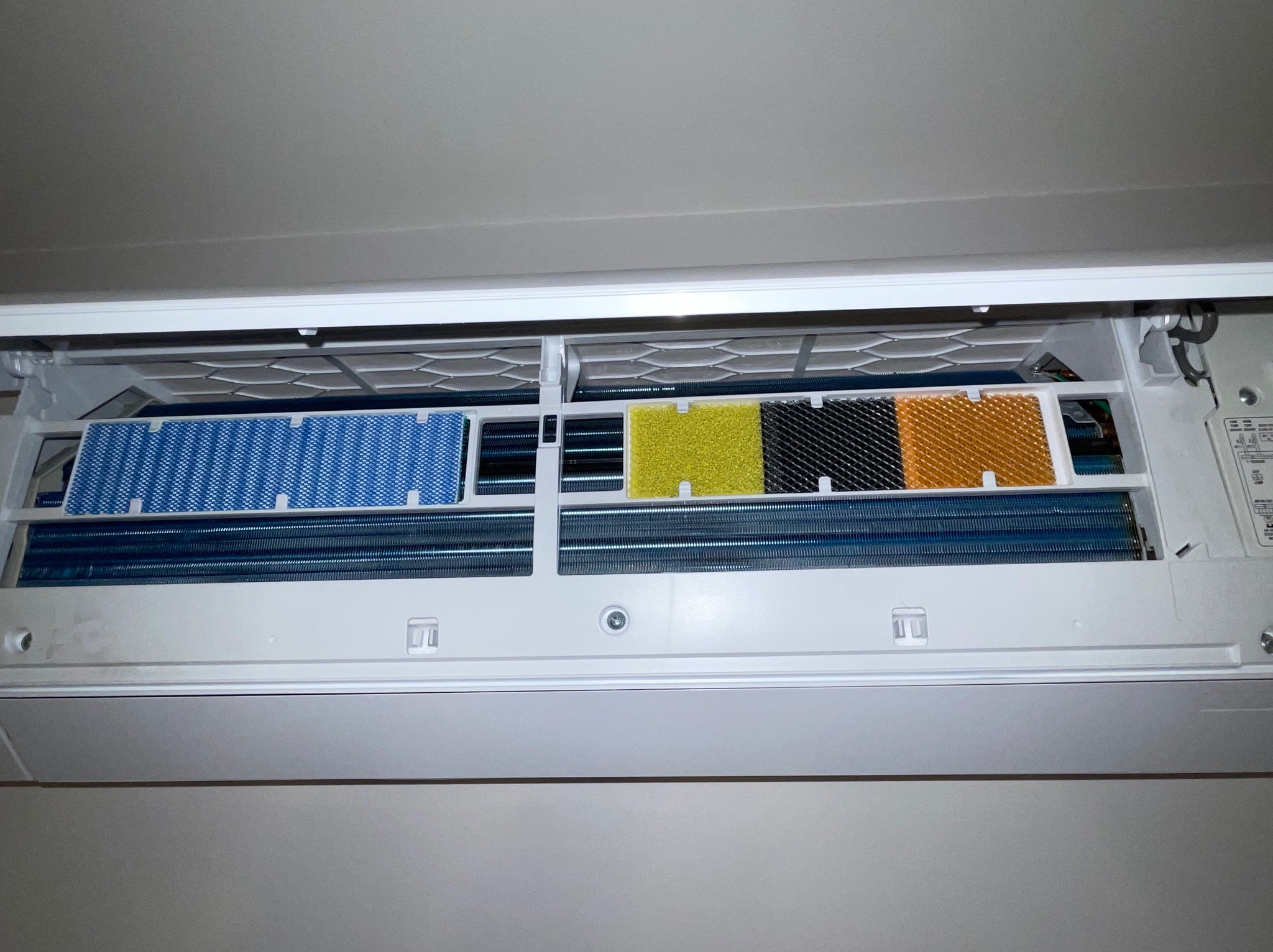 Otwarty klimatyzator ścienny z widocznymi filtrami w kolorach: niebieskim, żółtym, czarnym i pomarańczowym. Widoczne żebra chłodzące.