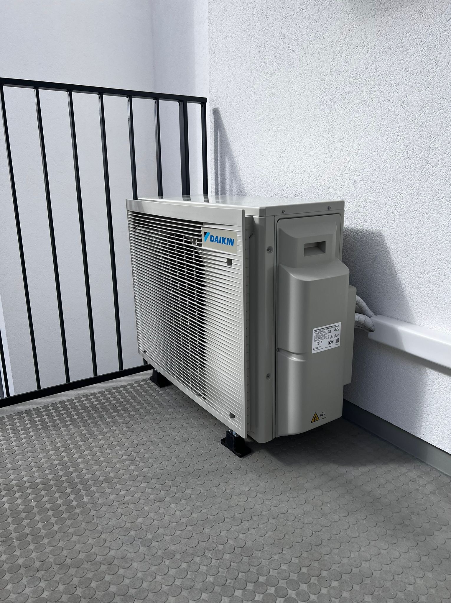 Zewnętrzna jednostka klimatyzacji Daikin na balkonie, widok z boku. Szara podłoga z okrągłymi wzorami, biała ściana z fakturą i czarna barierka w tle.