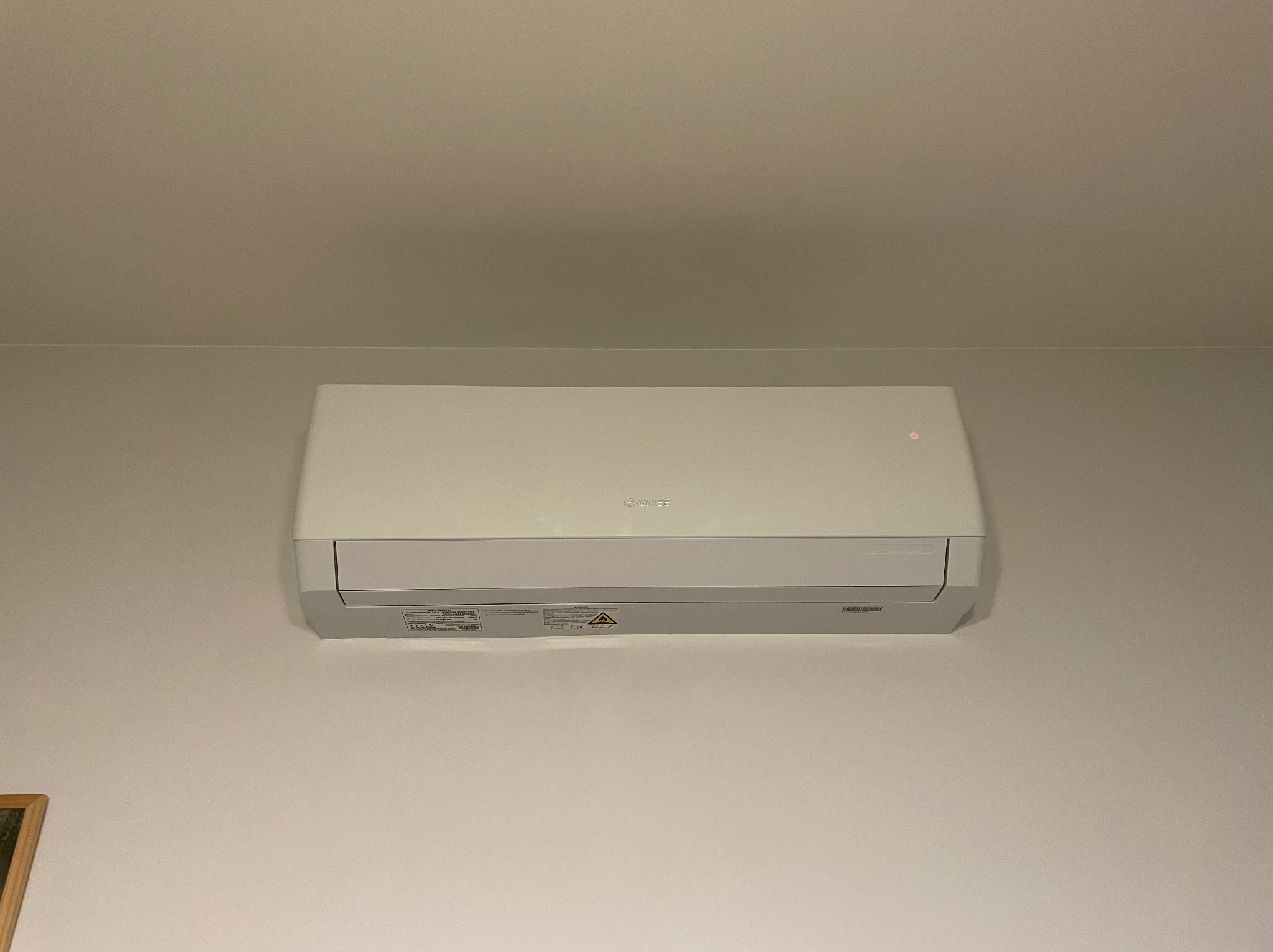 Biały klimatyzator marki Gree zamontowany na jasnej ścianie w pomieszczeniu. Widoczny z bliska, centralnie umieszczony na zdjęciu, z delikatnym oświetleniem.