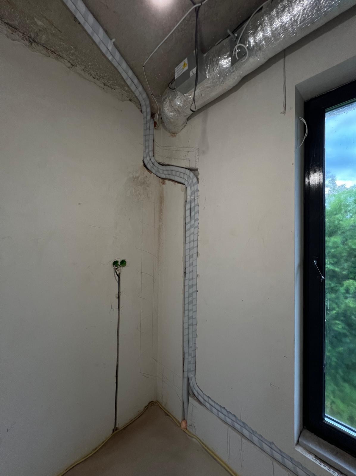 Montaż instalacji klimatyzacyjnej w surowym wnętrzu: rury w izolacji prowadzone po ścianach, widoczny kanał wentylacyjny i okno z zielonym widokiem na zewnątrz.