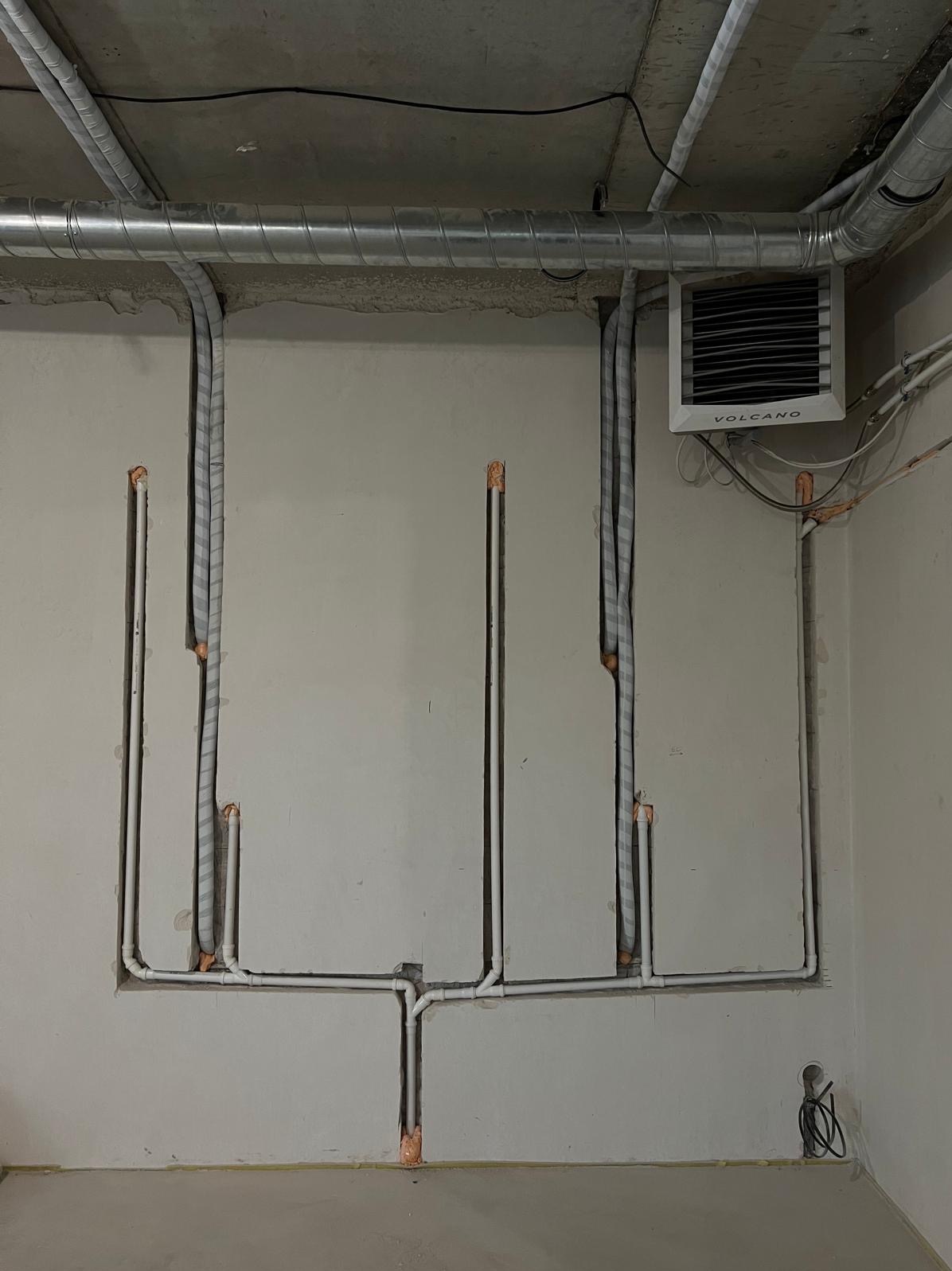 Instalacja grzewcza Volcano z rurami w ścianie, widoczne kanały i izolacja. Szare ściany w surowym stanie, rury prowadzone w bruzdach.
