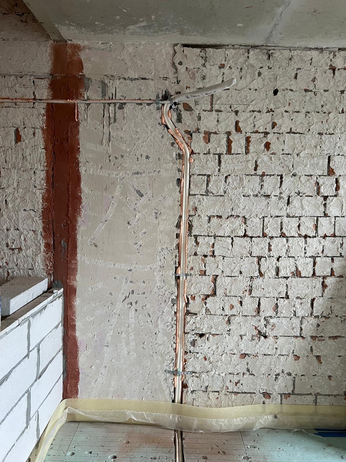 Montaż rur chłodniczych na surowej ścianie z cegły i tynku. Rury zabezpieczone pianką, przymocowane metalowymi uchwytami. Folia ochronna na podłodze.