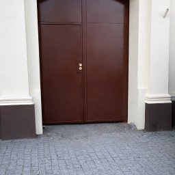 KAROL SZULAKIEWICZ - Dwuskrzydłowa, brązowa brama wjazdowa z łukowym nadprożem, osadzona w jasnej elewacji z numerem '6'. Brukowana nawierzchnia przed bramą. Minimalistyczny styl.