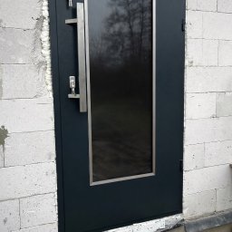 KAROL SZULAKIEWICZ - Ciemne, metalowe drzwi zewnętrzne z szybą i długim uchwytem, zamontowane w surowym murze z betonu komórkowego. Widoczne klucze w zamku.