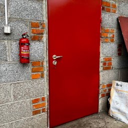 KAROL SZULAKIEWICZ - Czerwone, metalowe drzwi wejściowe z klamką i kluczem, zamontowane w szarej ścianie z ceglanymi wstawkami. Obok gaśnica i worek karmy dla zwierząt.