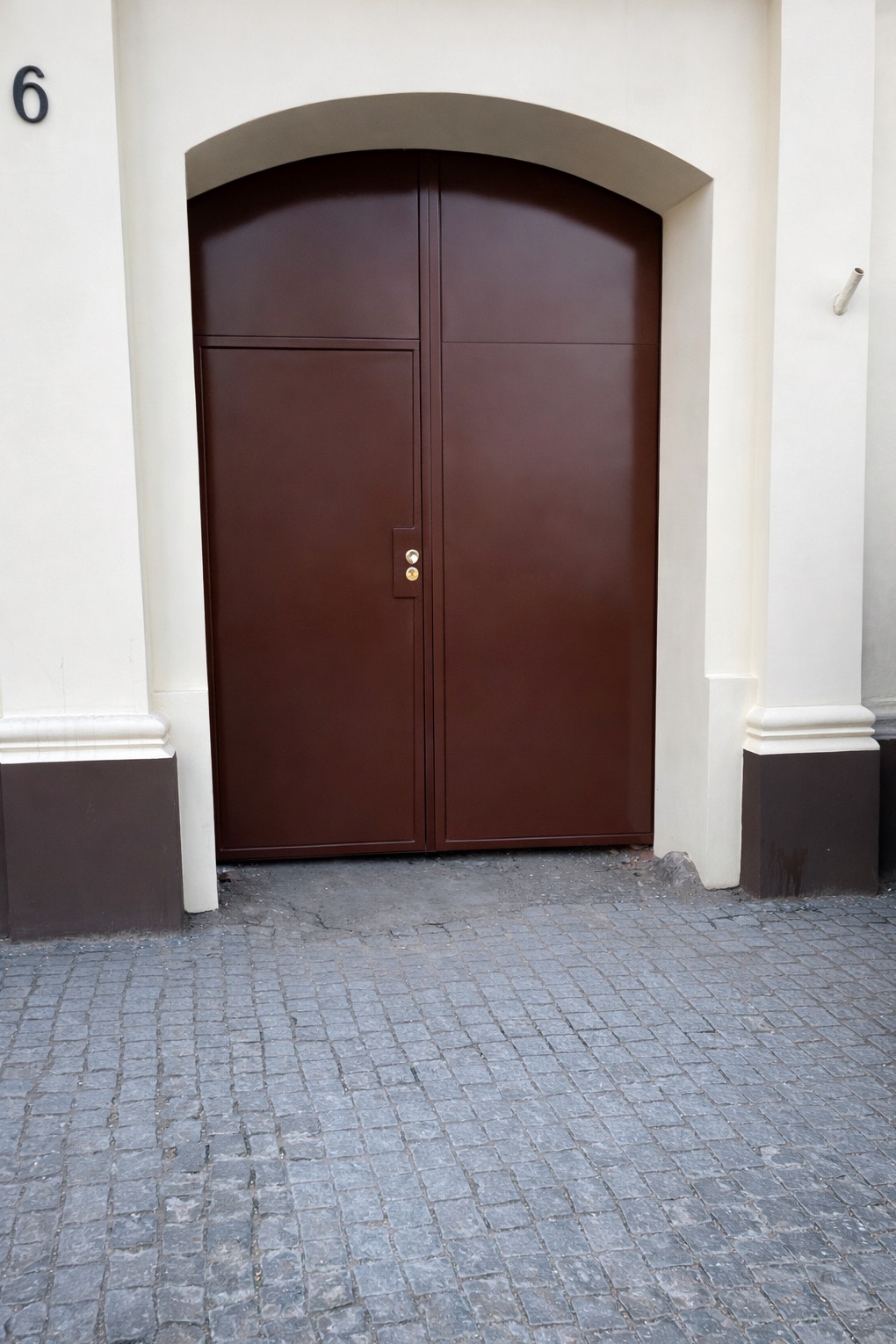 Dwuskrzydłowa, brązowa brama wjazdowa z łukowym nadprożem, osadzona w jasnej elewacji z numerem '6'. Brukowana nawierzchnia przed bramą. Minimalistyczny styl.