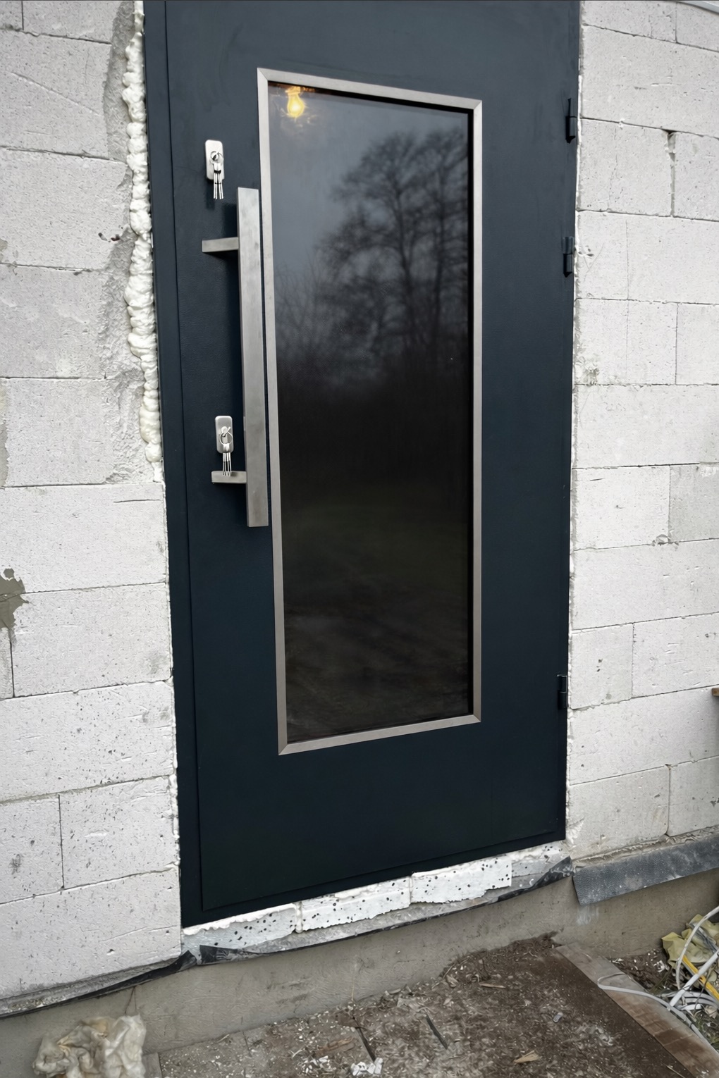 Ciemne, metalowe drzwi zewnętrzne z szybą i długim uchwytem, zamontowane w surowym murze z betonu komórkowego. Widoczne klucze w zamku.