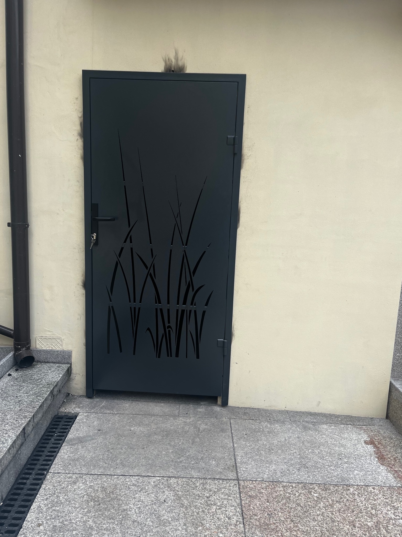 Metalowa furtka z wycinanym motywem roślinnym, umieszczona przy schodach i ścianie budynku. Ciemna kolorystyka kontrastuje z jasnym tłem.
