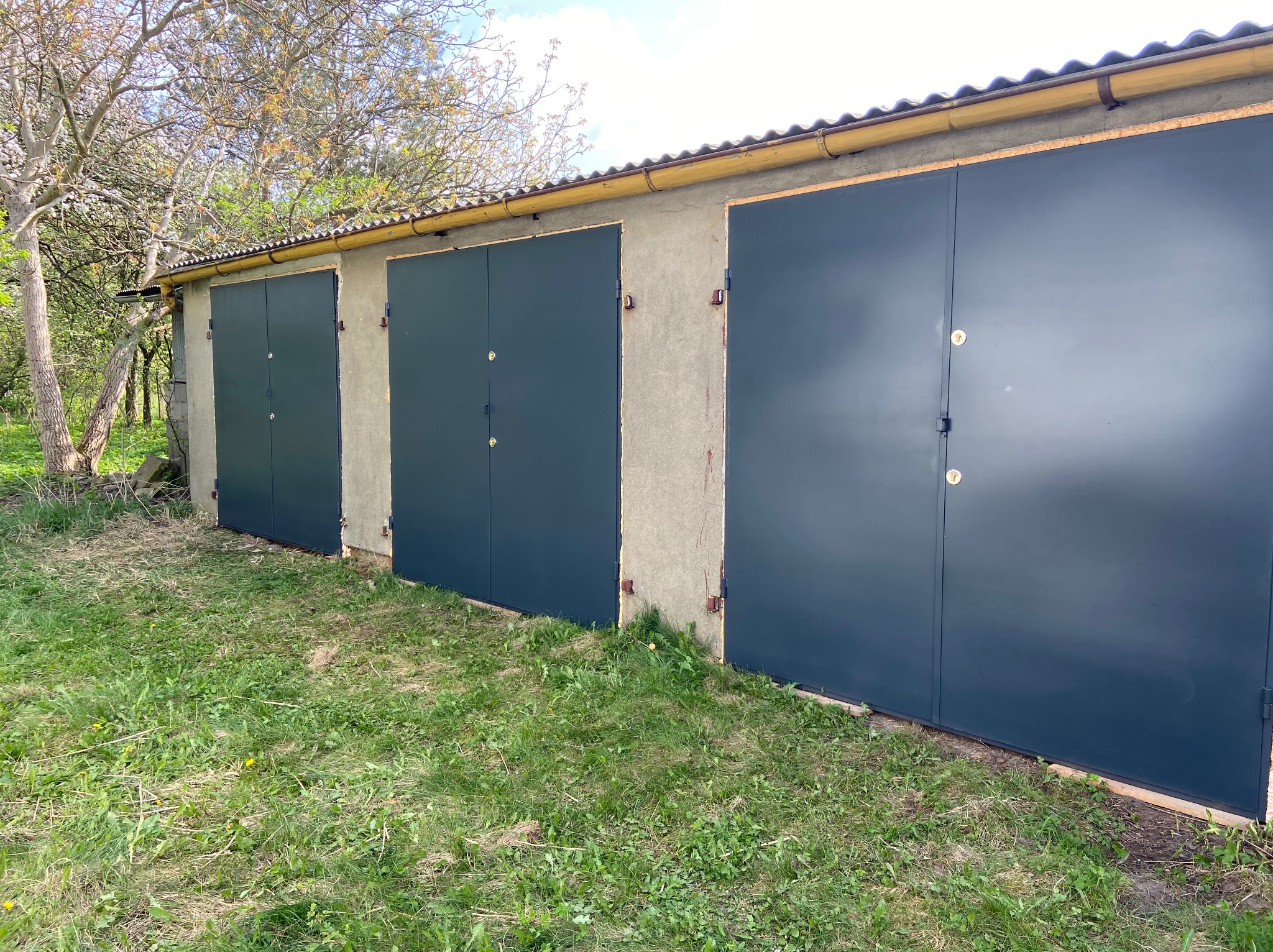 Trzy garaże z ciemnymi, metalowymi bramami segmentowymi, widoczne z zewnątrz na tle zielonej trawy i drzew. Brama po prawej stronie z widocznym zamkiem.