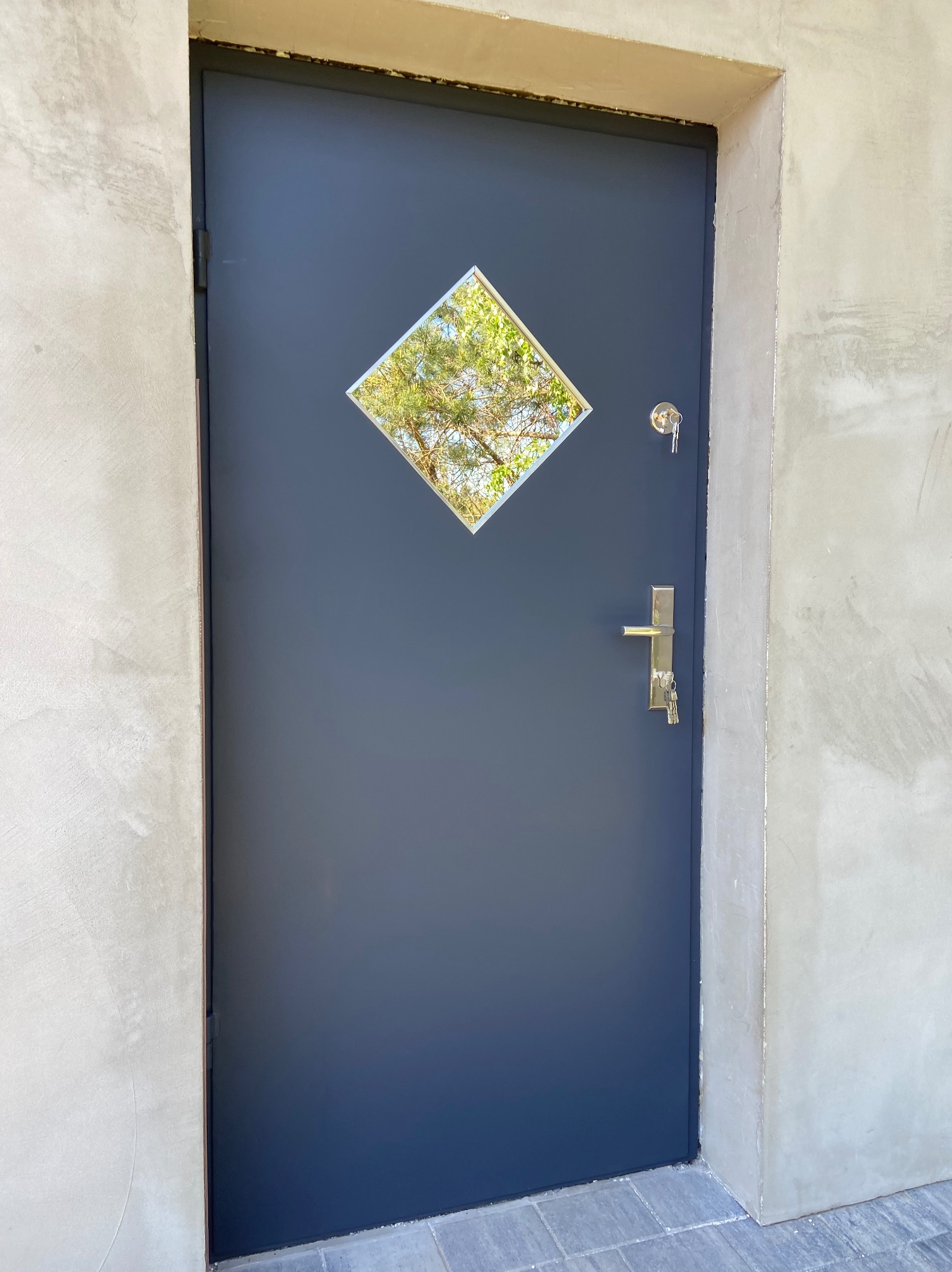 Ciemna, metalowa brama wejściowa z romboidalnym okienkiem, klucze w zamku, surowy beton wokół. Minimalistyczny design.