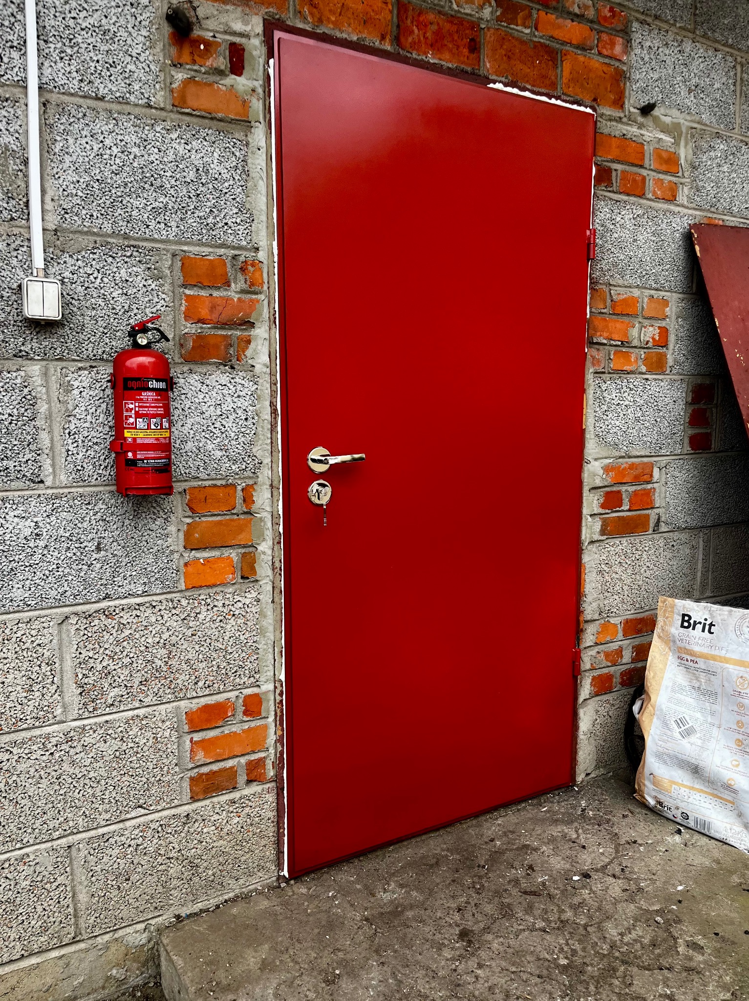 Czerwone, metalowe drzwi wejściowe z klamką i kluczem, zamontowane w szarej ścianie z ceglanymi wstawkami. Obok gaśnica i worek karmy dla zwierząt.