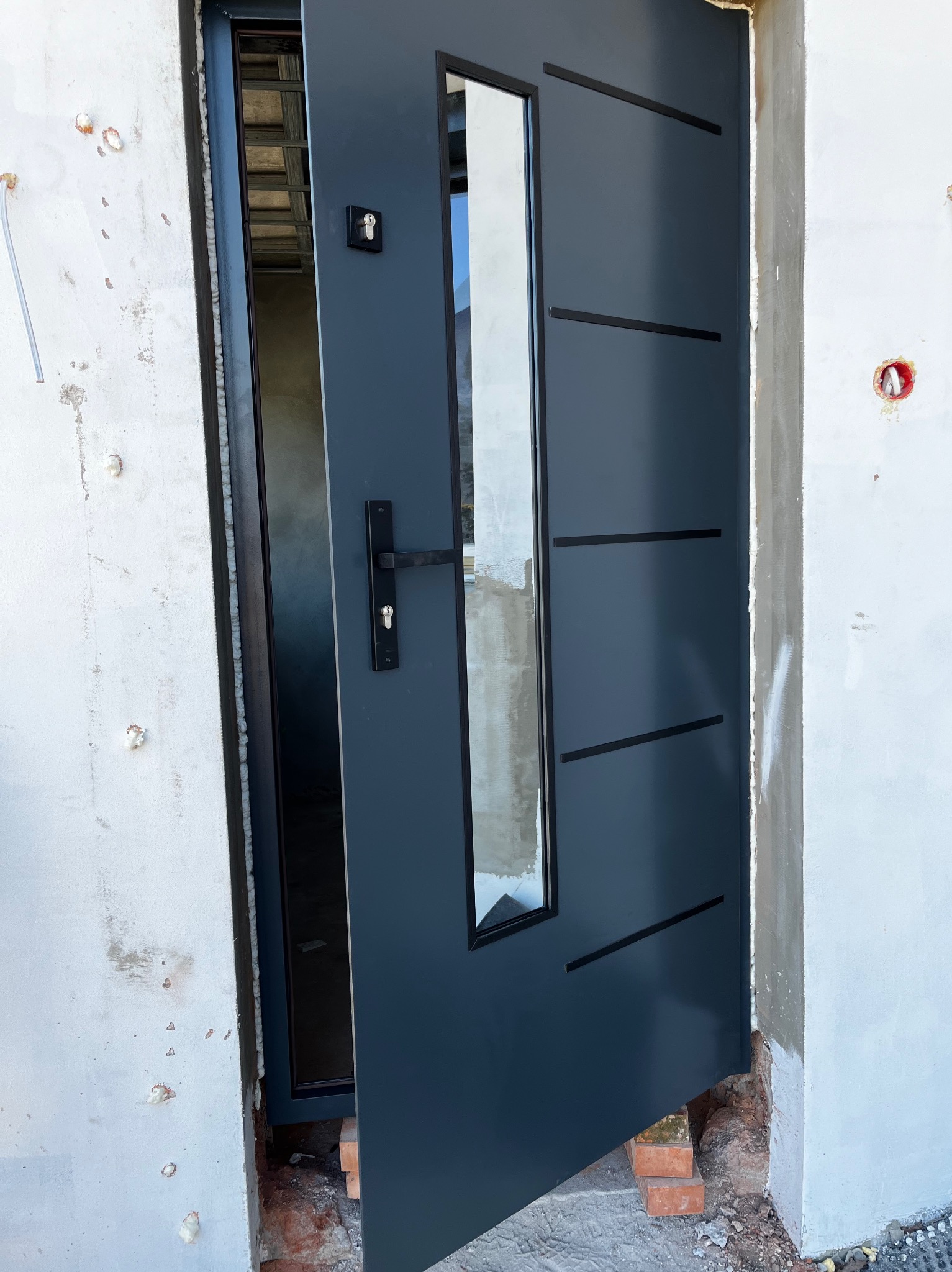 Grafitowe drzwi wejściowe z szybą, zamontowane w surowym otworze. Widoczne pianka montażowa i cegły pod progiem. Minimalistyczny design z poziomymi frezami.