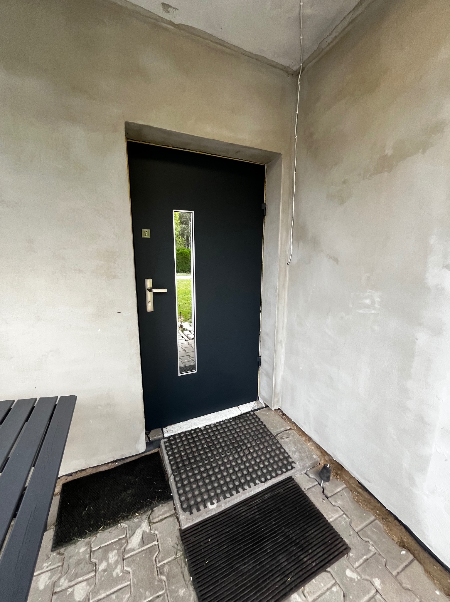 Ciemne drzwi wejściowe z przeszkleniem, zamontowane w surowym otoczeniu. Widoczne wycieraczki i fragment stołu. Ściany w stanie deweloperskim.