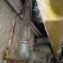 Bartosz Juszczak - Stara, skorodowana rura kanalizacyjna z widocznymi śladami rdzy na tle ściany pokrytej pianką izolacyjną i innymi rurami. Ujęcie z dołu, podkreślające zużycie instalacji.