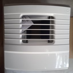 Bartosz Juszczak - Biały wentylator Aereco z kratką, widoczny z przodu, z instrukcją w środku i fragmentem kartonu obok, na neutralnym tle.