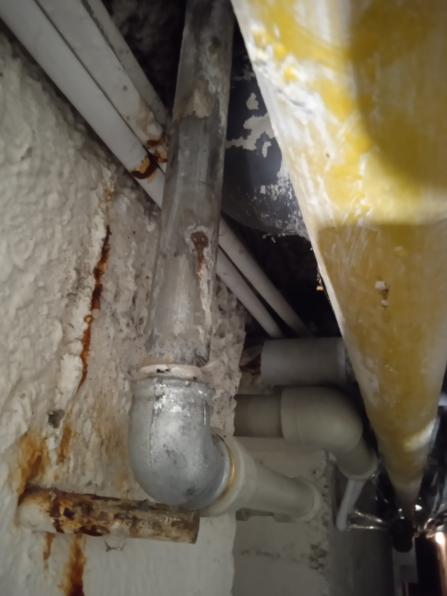 Stara, skorodowana rura kanalizacyjna z widocznymi śladami rdzy na tle ściany pokrytej pianką izolacyjną i innymi rurami. Ujęcie z dołu, podkreślające zużycie instalacji.