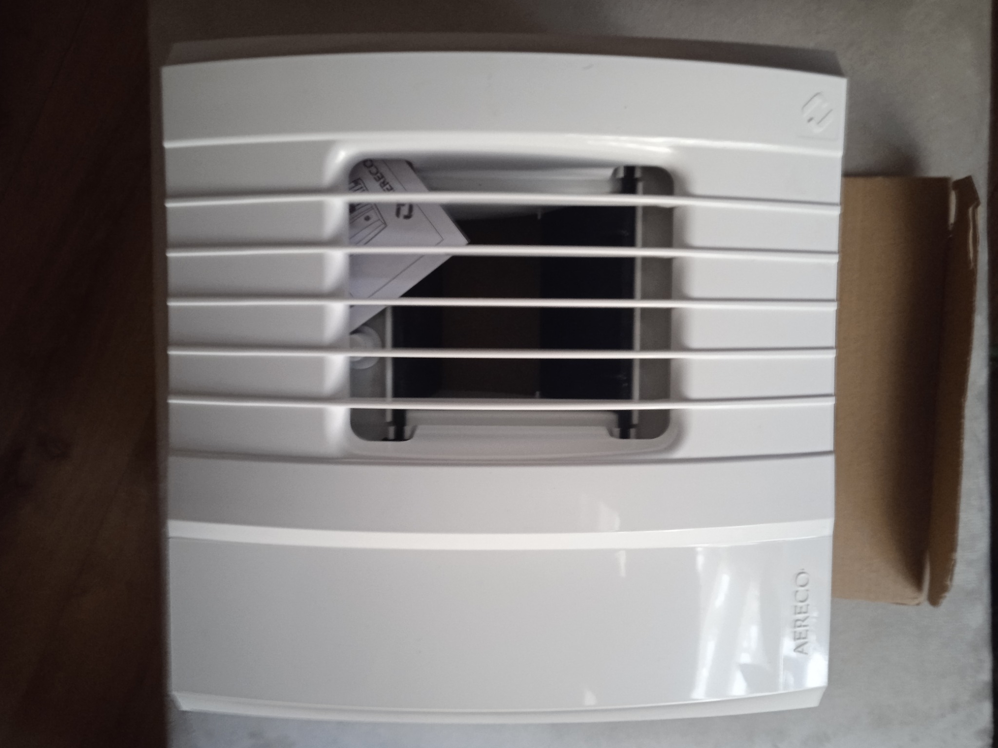 Biały wentylator Aereco z kratką, widoczny z przodu, z instrukcją w środku i fragmentem kartonu obok, na neutralnym tle.