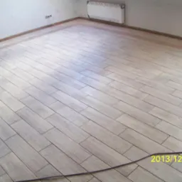 Ułożona podłoga z płytek imitujących drewno w jasnym odcieniu, widoczne listwy przypodłogowe i fragment grzejnika na ścianie. Widoczny kabel na podłodze.