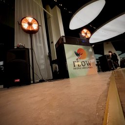 FLOW Iwona Smolińska - Scena z nagłośnieniem i oświetleniem, logo 'Flow Iwona Smolińska', reflektor na statywie, kolumny głośnikowe, dekoracyjne lampki. Ukośny kadr z perspektywą podłogi.