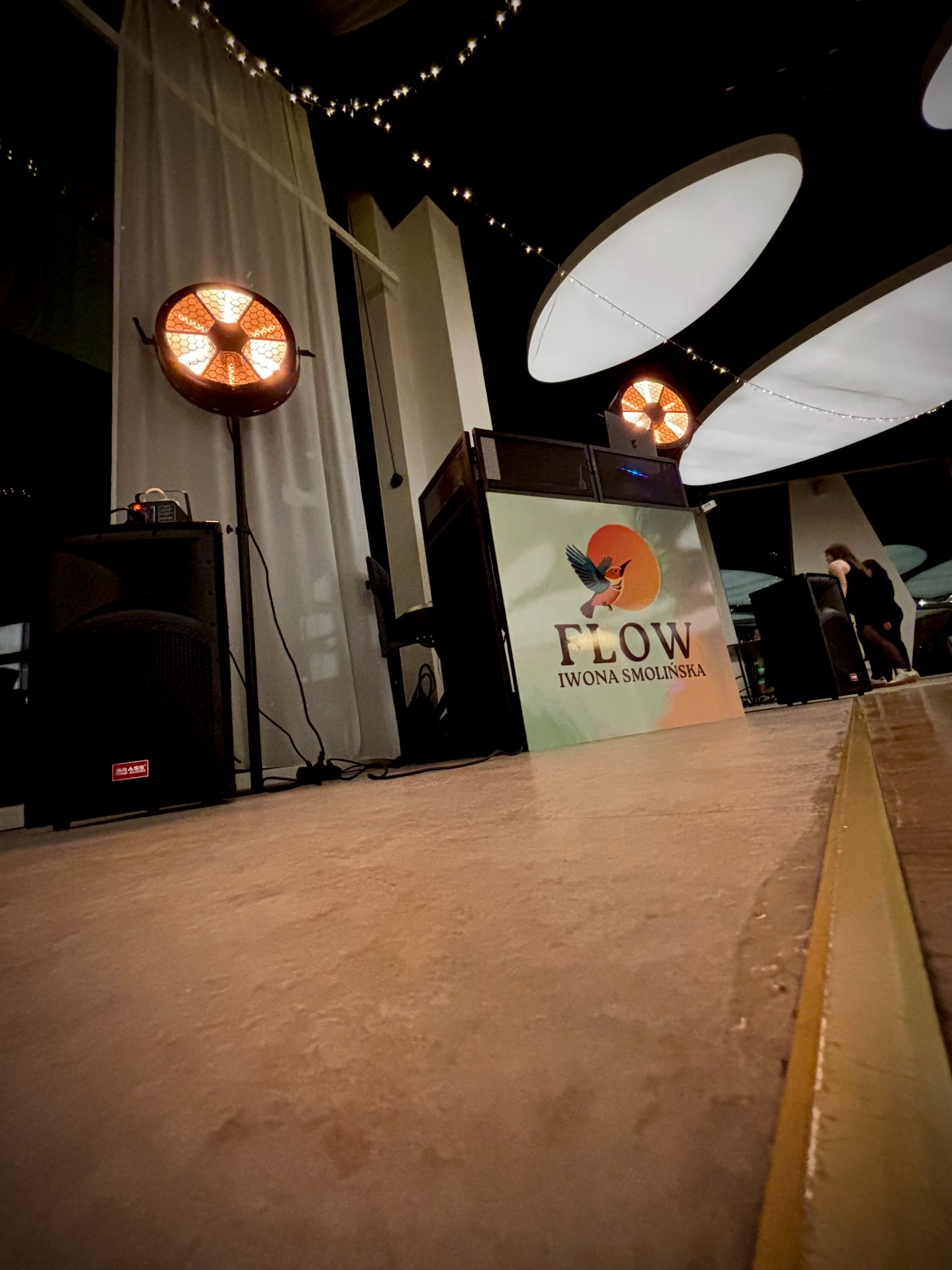 Scena z nagłośnieniem i oświetleniem, logo 'Flow Iwona Smolińska', reflektor na statywie, kolumny głośnikowe, dekoracyjne lampki. Ukośny kadr z perspektywą podłogi.