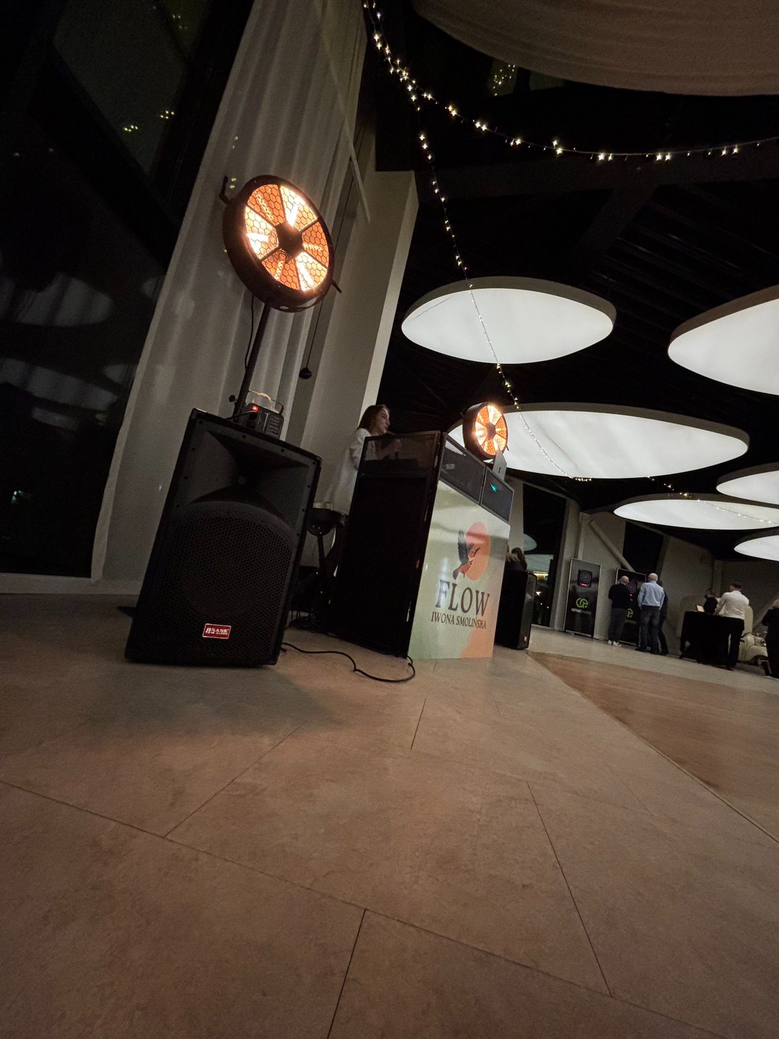 Sprzęt nagłośnieniowy i oświetleniowy na sali eventowej z DJ-ką. W tle widoczni goście. Charakterystyczne lampy grzewcze i dekoracyjne oświetlenie sufitowe.