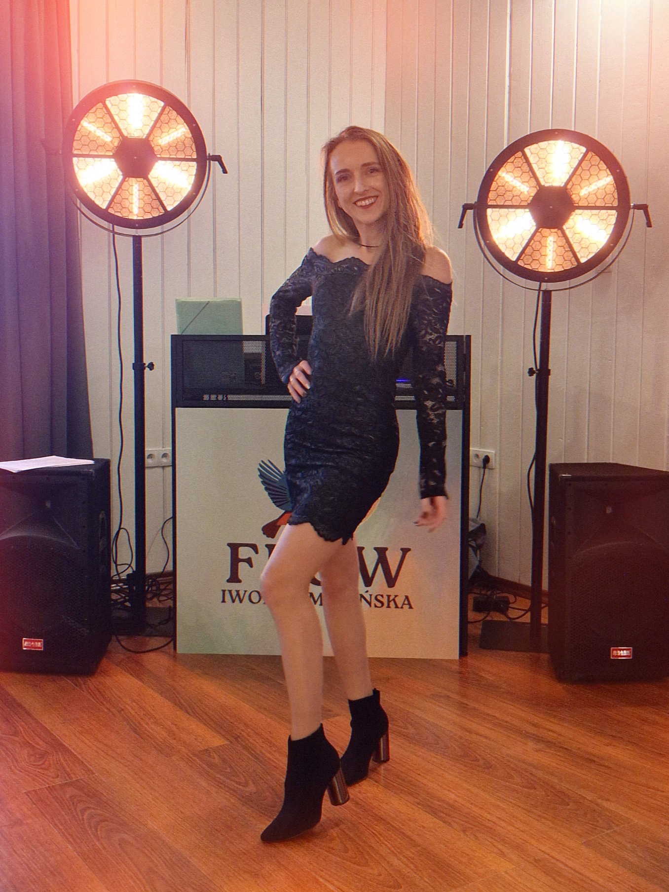 Uśmiechnięta kobieta w koronkowej sukience pozuje między dwoma reflektorami na statywach. W tle sprzęt muzyczny i logo firmy.