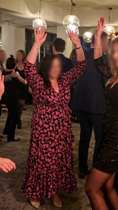 Rozbawiona kobieta w kwiecistej sukience tańczy z podniesionymi rękami na parkiecie podczas imprezy. W tle widoczne inne osoby i dekoracyjne lampy.