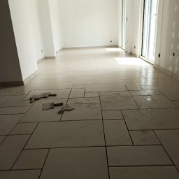 F.h.u. camry - Jasne, prostokątne płytki podłogowe ułożone w nowym, pustym pomieszczeniu. Widoczne narzędzia glazurnicze. Duże okno w tle. Świeżo położona podłoga.