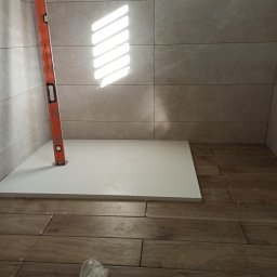 F.h.u. camry - Montaż brodzika w łazience z poziomnicą. Na podłodze i ścianach ułożone płytki imitujące drewno. Widoczne krzyżyki dystansowe.