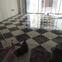 F.h.u. camry - Ukończona podłoga z czarno-białych płytek ułożonych w karo, widoczne narzędzia i akcesoria glazurnicze, ściany przygotowane do dalszych prac wykończeniowych.