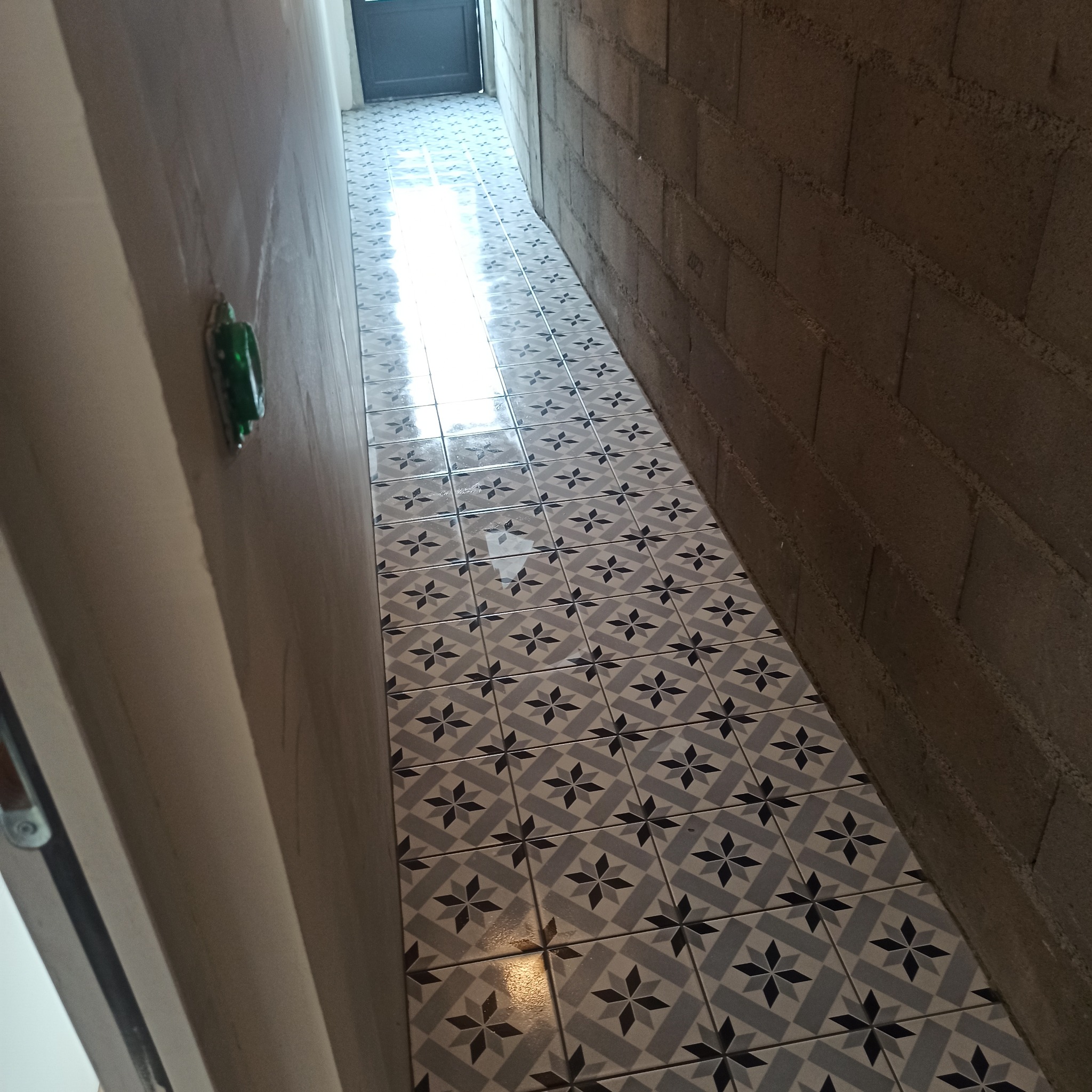 Wąski korytarz z nową, wzorzystą podłogą z płytek w odcieniach szarości i bieli. Ściana z surowych betonowych bloków po prawej, gładka, jasna ściana po lewej.