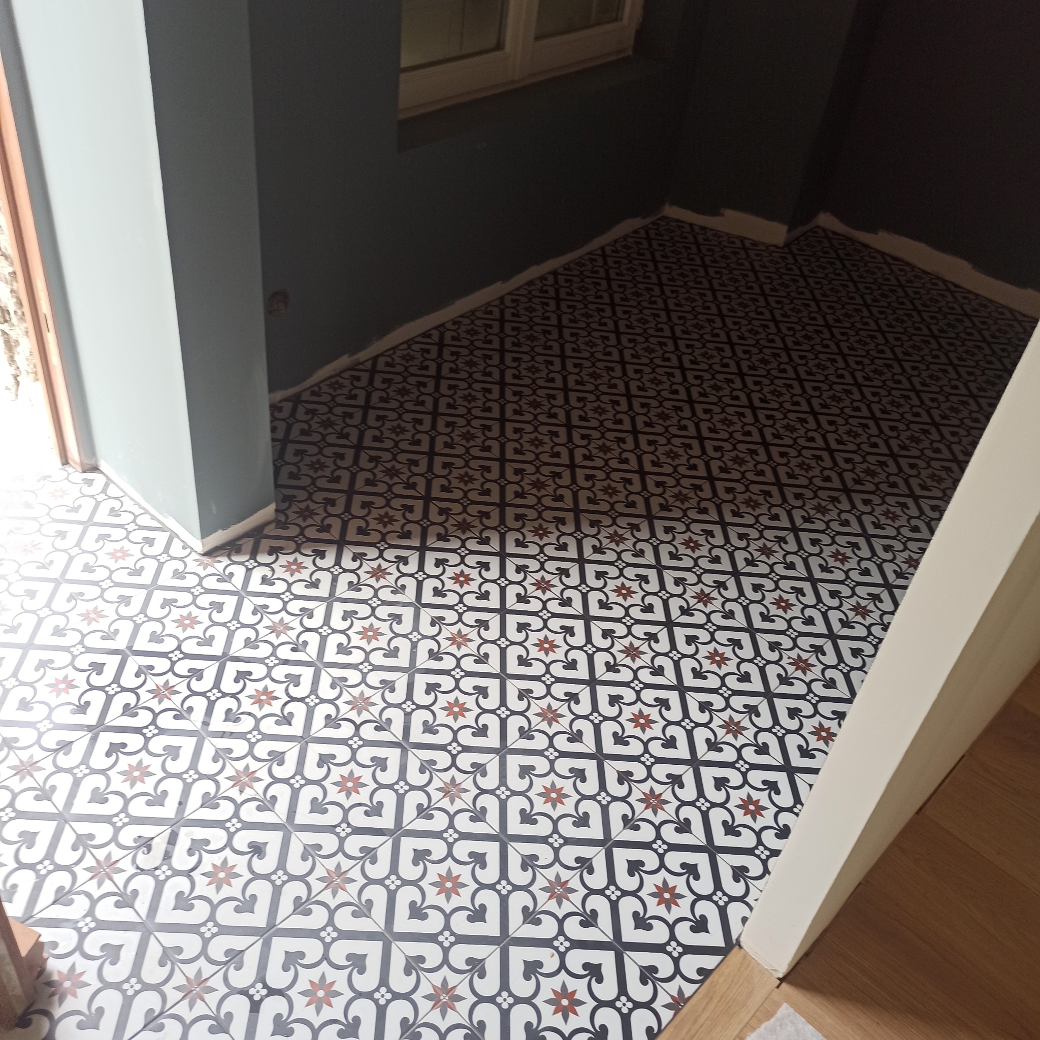Wzorzysta, nowa podłoga z płytek w odcieniach bieli, czerni i czerwieni, ułożona w pomieszczeniu z ciemnymi ścianami i oknem. Widoczny fragment progu i schodów.
