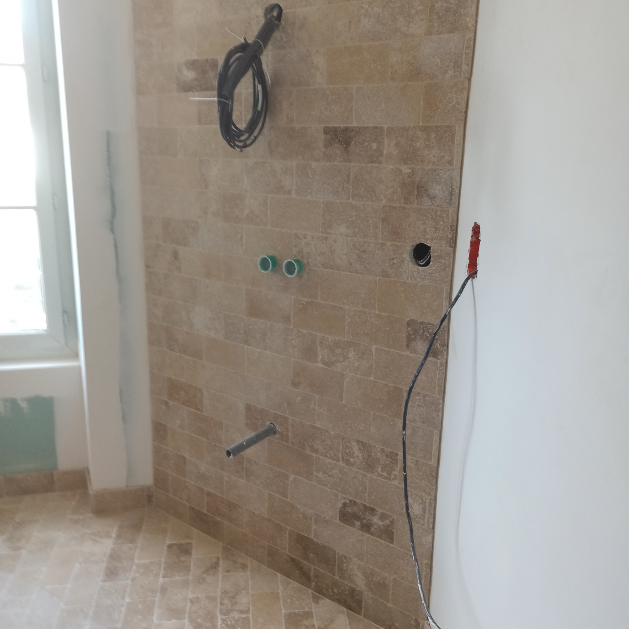 Ściana łazienki wyłożona beżową cegłą, z widocznymi instalacjami wodnymi i elektrycznymi. W tle okno i fragment białej ściany. Widoczne niedokończone prace.