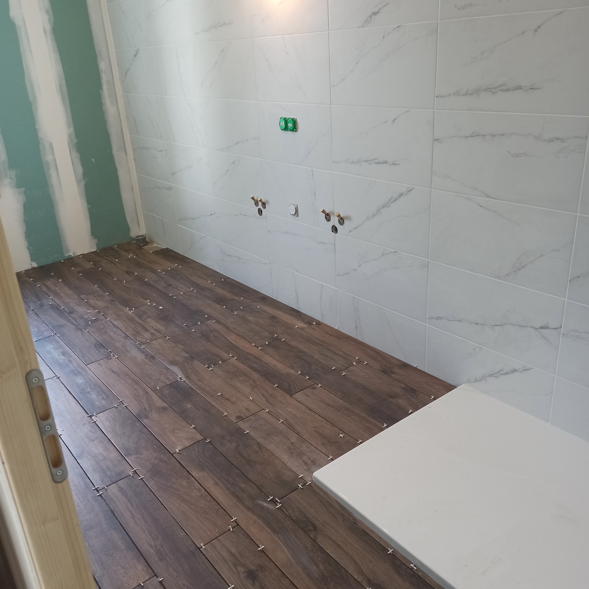 Łazienka w trakcie prac: ułożona podłoga z płytek imitujących drewno, białe płytki na ścianach z marmurkowym wzorem, widoczne elementy instalacji i płyta gipsowa.